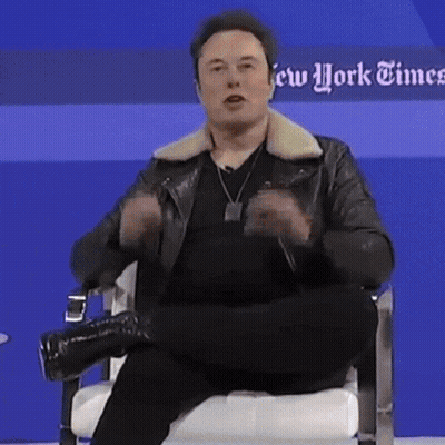 A Man In A Leather Jacket Is Sitting In A Chair In Front Of A Blue Wall That Says New York Times GIF