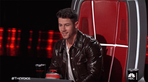 A Man In A Leather Jacket Is Sitting In A Chair With A Nbc Logo On The Back GIF