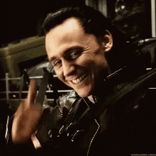 A Man In A Leather Jacket Is Smiling And Waving While Holding A Gun . GIF