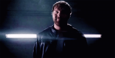A Man In A Leather Jacket Is Standing In A Dark Room With A Light Shining On Him . GIF