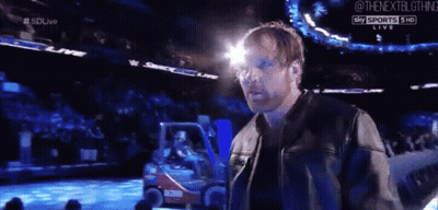 A Man In A Leather Jacket Is Standing In Front Of A Crowd At A Wrestling Event . GIF