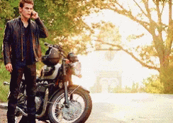 A Man In A Leather Jacket Is Standing Next To A Motorcycle Talking On A Cell Phone GIF