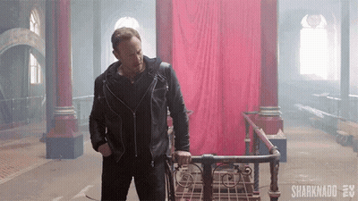 A Man In A Leather Jacket Is Standing Next To A Walker In A Room With A Red Curtain Behind Him . GIF