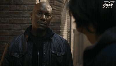 A Man In A Leather Jacket Is Talking To Another Man In Front Of A Brick Wall With X Fast Written On It GIF