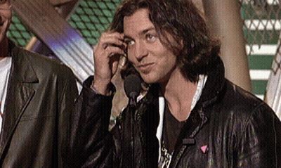 A Man In A Leather Jacket Talks Into A Microphone While Another Man Stands Behind Him GIF