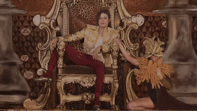 A Man In A Leopard Print Jacket Is Sitting On A Throne With A Woman Behind Him GIF