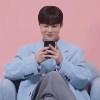 A Man In A Light Blue Coat Is Sitting In A Chair Looking At His Phone GIF