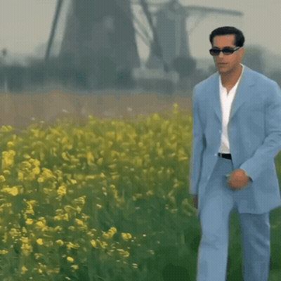 A Man In A Light Blue Suit And Sunglasses Walks Through A Field Of Yellow Flowers GIF