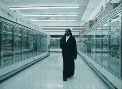 A Man In A Long Black Coat Walks Through An Empty Supermarket Aisle GIF