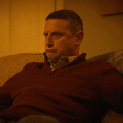 A Man In A Maroon Sweater And Tie Sits On A Couch GIF