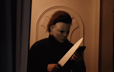 A Man In A Mask Is Holding A Knife And Looking At His Phone GIF