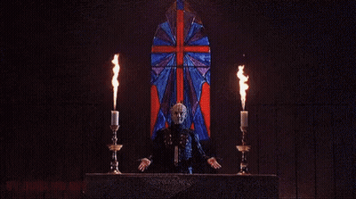 A Man In A Mask Is Holding Two Candles In Front Of A Burning Building . GIF