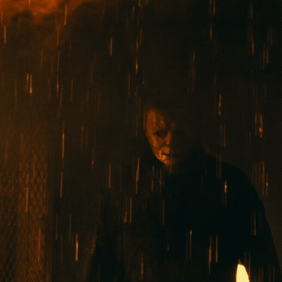 A Man In A Mask Is Standing In The Rain With A Fire In The Background GIF
