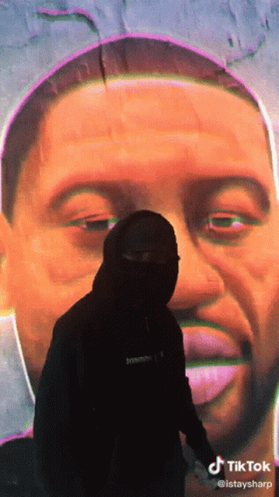 A Man In A Mask Stands In Front Of A Large Painting Of A Man 'S Face GIF