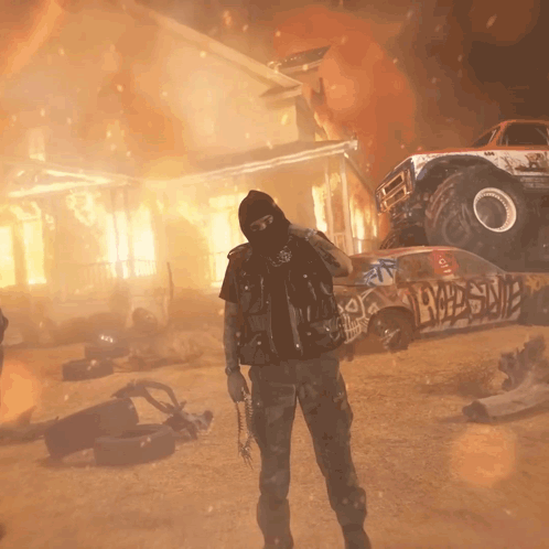 A Man In A Mask Stands In Front Of A Monster Truck And A Burning House With Graffiti On It GIF