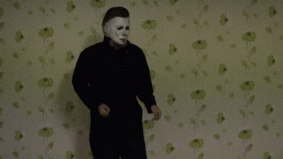 A Man In A Mask Stands In Front Of A Wall With Green Flowers On It GIF