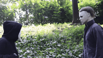 A Man In A Mask Stands Next To Another Man In A Hood GIF