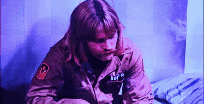 A Man In A Military Jacket With The Word Bof On His Chest Is Sitting On A Bed . GIF