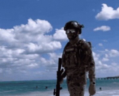 A Man In A Military Uniform Is Standing On A Beach Holding A Gun GIF