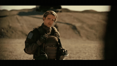 A Man In A Military Uniform Stands In A Desert Holding A Helmet GIF