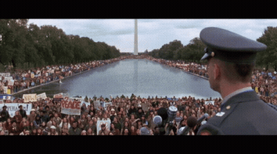 A Man In A Military Uniform Stands In Front Of A Crowd Holding Signs Including One That Says " Peace " GIF