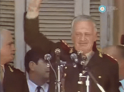 A Man In A Military Uniform Stands In Front Of Two Microphones And Waves His Hand GIF
