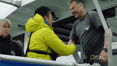 A Man In A National Geographic Shirt Shakes Hands With Another Man On A Boat GIF
