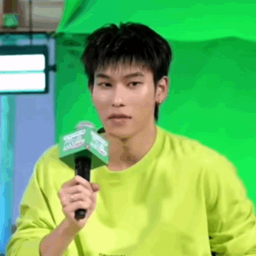 A Man In A Neon Green Shirt Is Holding A Microphone In Front Of A Green Screen . GIF