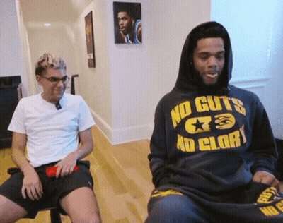 A Man In A No Guts No Glory Hoodie Sits Next To Another Man GIF