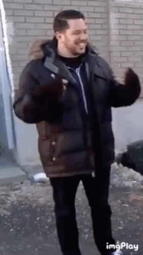 A Man In A Parka Is Standing In Front Of A Brick Wall And Waving . GIF