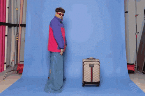 A Man In A Pink And Purple Jacket Is Standing Next To A Suitcase GIF