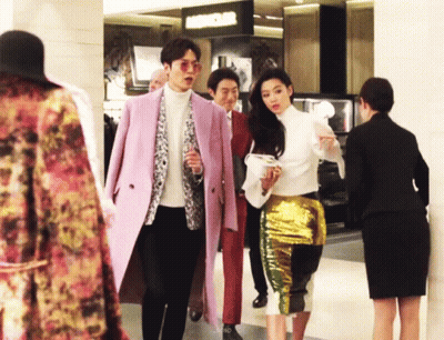 A Man In A Pink Coat And A Woman In A Gold Skirt Are Walking In A Store GIF