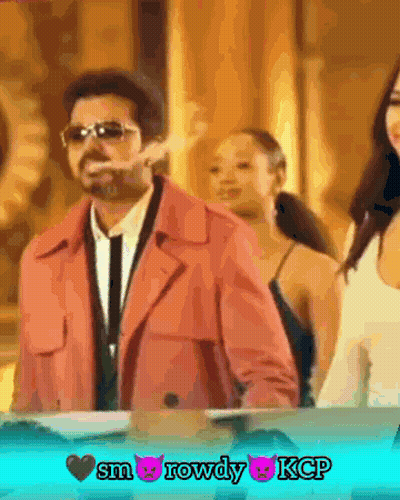 A Man In A Pink Coat Is Smoking A Cigarette In Front Of Two Women GIF