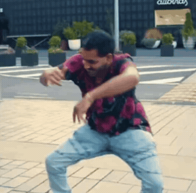 A Man In A Pink Shirt And Blue Jeans Is Dancing On A Brick Sidewalk . GIF