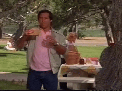 A Man In A Pink Shirt Is Eating A Hamburger In The Park . GIF
