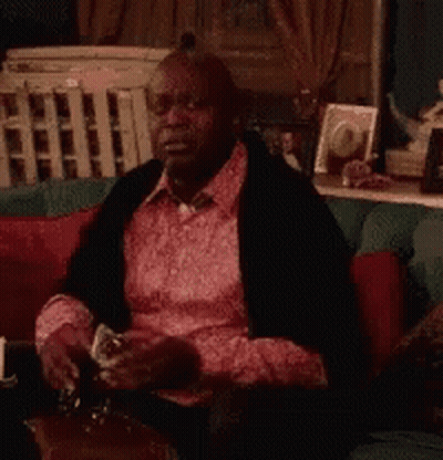 A Man In A Pink Shirt Is Sitting On A Couch With His Hands Up . GIF