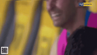 A Man In A Pink Shirt Is Smiling In Front Of A Tico Sports Sign GIF