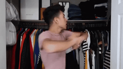 A Man In A Pink Shirt Is Standing In A Closet Filled With Clothes And Bags . GIF