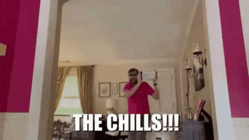 A Man In A Pink Shirt Is Standing In A Living Room With The Words The Chills Written On The Wall Behind Him . GIF