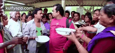 A Man In A Pink Shirt Is Standing In Front Of A Crowd Of People Holding Plates . GIF
