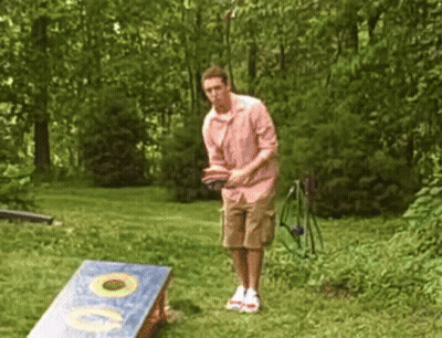 A Man In A Pink Shirt Is Throwing A Frisbee In A Cornhole Board GIF