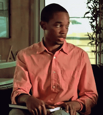 A Man In A Pink Shirt Sits In A Chair Holding A Book GIF