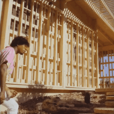 A Man In A Pink Shirt Stands In Front Of A Building With A Wooden Fence GIF