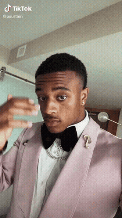A Man In A Pink Suit And Black Bow Tie Is Taking A Selfie . GIF
