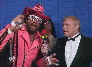 A Man In A Pink Suit Is Holding A Microphone While Talking To A Man In A Tuxedo . GIF