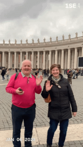 A Man In A Pink Sweater And A Woman In A Black Coat Are Standing In Front Of A Building With The Date Jan 06 2023 GIF
