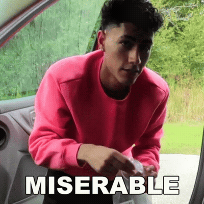a man in a pink sweater is standing in the doorway of a car with the word miserable written on the side .