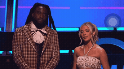 A Man In A Plaid Jacket Stands Next To A Woman In A White Crop Top GIF
