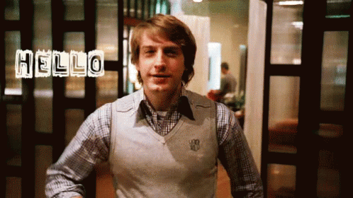 A Man In A Plaid Shirt And A White Vest Says Hello GIF