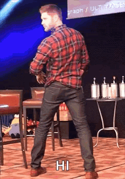 A Man In A Plaid Shirt And Black Jeans Is Standing On A Stage With His Hands In His Pockets . GIF
